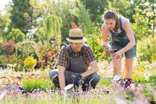 In a well-maintained front garden in Neasden, two women are engaged in gardening activities amidst vibrant flower beds. One woman, wearing a straw hat, a checkered shirt, and gardening gloves, is crouched down near a patch of flowering plants, possibly pruning or planting. The other woman, dressed in a sleeveless top and denim overalls, is leaning forward, holding garden shears and tending to nearby plants. The garden features a neatly trimmed lawn with dense green grass, bordered by rich soil and flower beds filled with a variety of colourful blooms. In the background, there are lush trees and shrubs providing natural privacy and shade, with dappled sunlight casting gentle shadows. The scene captures a bright and pleasant weather day, typical of outdoor gardening work in London, with clear lighting highlighting the textures of the garden elements. The image showcases a thriving outdoor space maintained by local gardening services such as Landscapers Neasden, reflecting a well-organized and inviting landscaped yard that emphasizes healthy plants, lawn care, and garden maintenance work.