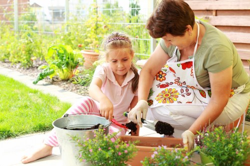 A woman and a young girl are engaged in gardening activities in a well-maintained outdoor garden space. The woman is wearing a floral apron and gardening gloves, actively planting or tending to the soil in a rectangular flower bed, which contains various green plants. The girl, seated on the grass, is wearing a light pink top with striped pants and is assisting with the gardening tasks. Surrounding them are vibrant green plants, some potted flowers, and a lush lawn with neatly trimmed grass. In the background, there are garden beds with leafy plants, a wire fence, and a wooden structure, likely a shed or garden wall. The scene suggests a sunny day with natural lighting, creating a warm and inviting outdoor environment perfect for gardening and outdoor maintenance, typical of landscaping and planting services in the local Neasden area, as featured on landscapersneasden.co.uk.
