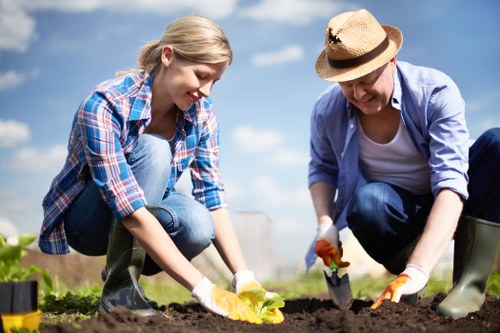 A woman and a man are working together in a garden, planting or tending to the soil with small gardening tools, both wearing casual clothing and gloves, amidst a lush green outdoor setting with a blue sky overhead. The foreground features freshly turned soil with a yellow flower in the woman's hand, while the background includes patches of grass, small plants, and garden tools scattered around, indicating a well-maintained landscaped outdoor space typical of residential gardens near Neasden or the London area. The scene depicts during daytime with natural sunlight illuminating the scene, embodying outdoor gardening activities that a professional landscaping company like Landscapers Neasden might support through garden and lawn care services.