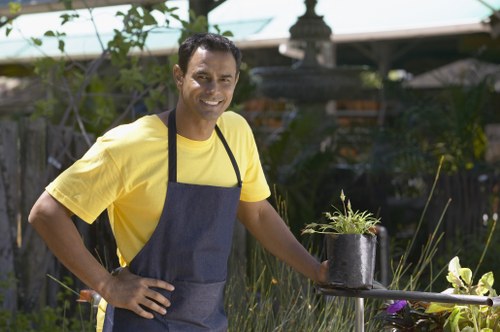 A male gardener with light brown skin, short dark hair, and a friendly expression, standing outdoors in a well-maintained garden during daytime. He is wearing a bright yellow T-shirt and a dark grey apron, and is holding a potted plant with green foliage in his right hand while resting his left hand on his hip. The background features a lush garden with tall plants, bushes, and trees, along with a wooden fence and a greenhouse structure partially visible. The garden area has a mix of flower beds, shrubbery, and a paved surface, with natural sunlight illuminating the scene, reflecting a tidy and cultivated outdoor space suitable for professional gardening and landscaping services in the Neasden area, nearby London.