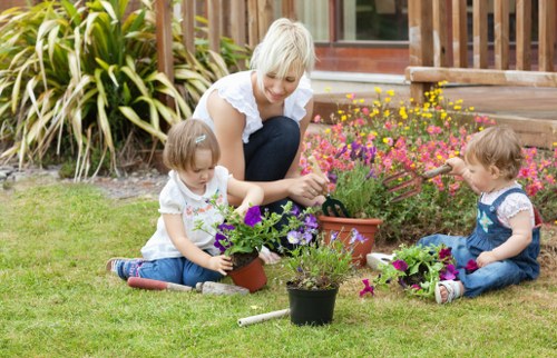 A woman with short blonde hair and two young children are working together in a well-maintained front garden, tending to flower pots and a flower bed near a wooden deck. The garden features a lush, green lawn with dense, ornamental foliage and flowering plants in shades of pink, purple, and yellow, which are planted in borders and containers. The children, dressed in casual clothes, are engaged in planting and watering, while the woman assists and supervises. The scene takes place during daytime with natural lighting, highlighting the vibrant colours and textures of the garden. In the background, a wooden deck, part of a residential property in Neasden, provides a backdrop for outdoor family activity and gardening. The overall setting emphasizes a tidy, inviting outdoor space with a focus on gardening activities suitable for a family-friendly environment, supporting outdoor landscaping and lawn care services offered by Landscapers Neasden.