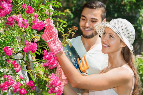 A man and woman are using gardening tools and wearing protective gloves while tending to a vibrant pink flowering shrub in a well-maintained outdoor garden. The woman, wearing a wide-brimmed, polka-dotted sun hat, is gently pruning the flowers with pruning shears, while the man, with short dark hair and a beard, is smiling and observing her work. The garden features lush green foliage, including dense shrubbery and trees in the background, with a mix of flowering plants and leafy plants creating a natural, colourful environment. The scene is set under bright daylight, highlighting the natural tones of the plants and the freshly cut flowers, indicating a pleasant outdoor gardening activity in a residential setting. This image exemplifies outdoor garden maintenance and landscaping work typical of professional garden services in Neasden or the surrounding NW postcode area, as part of the landscaping and gardening services offered by Landscapers Neasden on their landscape gardening page.