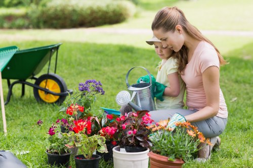 In a lush, well-maintained garden in Neasden, two young girls are engaged in gardening activities, surrounded by an array of colorful flowering plants in various pots and containers. The garden features a neatly mown grass lawn with vibrant green hues, bordered by a mixture of flowering plants with red, purple, pink, and orange blossoms, creating a lively and inviting outdoor space. In the background, a hedge and trees provide natural boundary and shade, while a green wheelbarrow with a yellow wheel is parked nearby, suggesting ongoing outdoor maintenance or planting work by locals or professional landscapers like Landscapers Neasden. The scene is set on a bright, sunny day with natural lighting highlighting the textured leaves, flower petals, and lush foliage, emphasizing an outdoor environment perfect for family gardening or professional landscaping services in the Neasden area. The girls are wearing casual clothing suitable for gardening, with one holding a small watering can, reinforcing the theme of garden care and outdoor activity in a residential setting.
