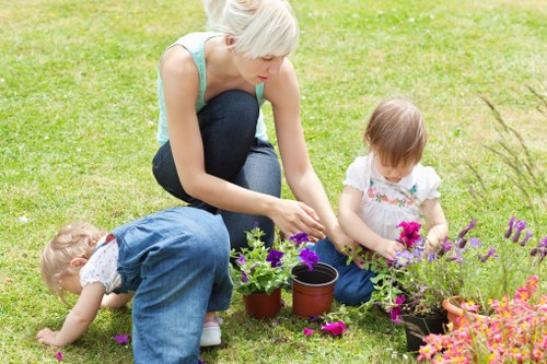 A woman with short blonde hair and a light blue sleeveless top is kneeling on a well-maintained grassy lawn in a garden, engaged in planting flowers. She is placing a potted plant into the soil while two young children, a girl and a boy, are nearby. The girl, wearing a white top with pink accents, is sitting on the grass holding gardening tools and examining a cluster of pink and purple flowers in a pot, while the boy, dressed in denim overalls and a white shirt, is crouched down, touching the grass or soil. The garden features a lush, evenly cut lawn with a border of vibrant flowering plants in various shades of pink, purple, and orange, creating a colourful, inviting outdoor space. In the background, there are some trees and shrubs, indicating a landscaped garden area. The weather appears clear, with bright natural light illuminating the scene. This image reflects typical outdoor gardening activities and the creation of a lively, colourful garden space, aligning with professional gardening and planting services offered by companies like Landscapers Neasden in the local area of Neasden, London, captured on the page titled 'Neasden Planting Services Guide'.